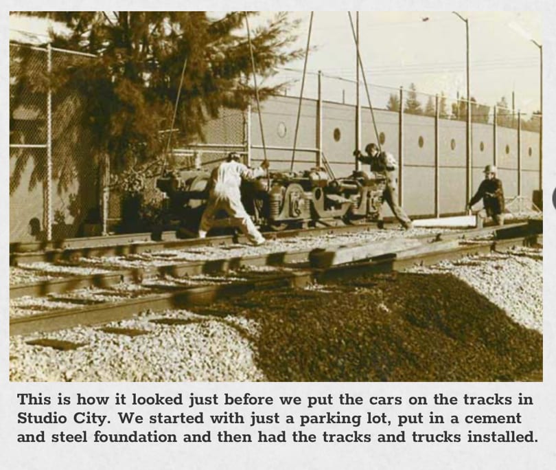 Three workers installing train trucks onto tracks in Studio City, with a fence and building in the background.