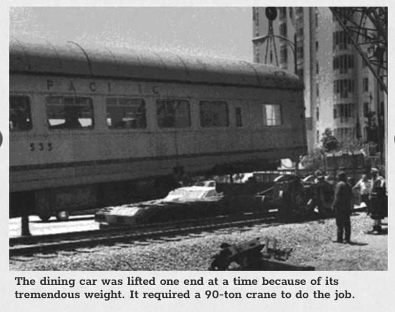 Black and white photo of a crane lifting a "Pacific" train car, with people watching nearby.