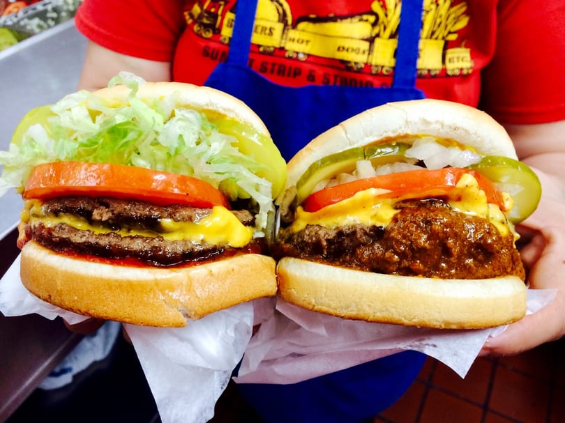 Two hands hold a double cheeseburger with lettuce and tomato, and a chili cheeseburger with pickles.