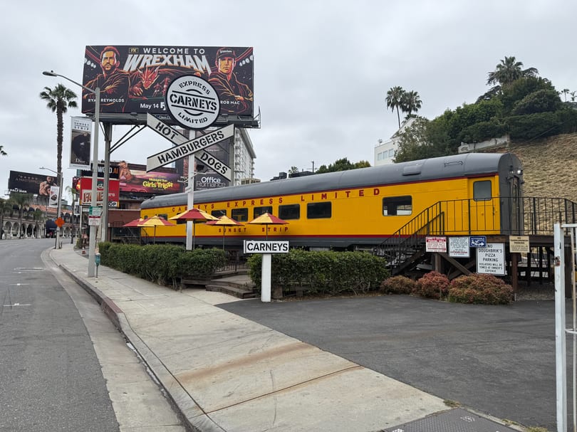 Carney's restaurant, a yellow train car with red umbrellas, sits beside a street with billboards.
